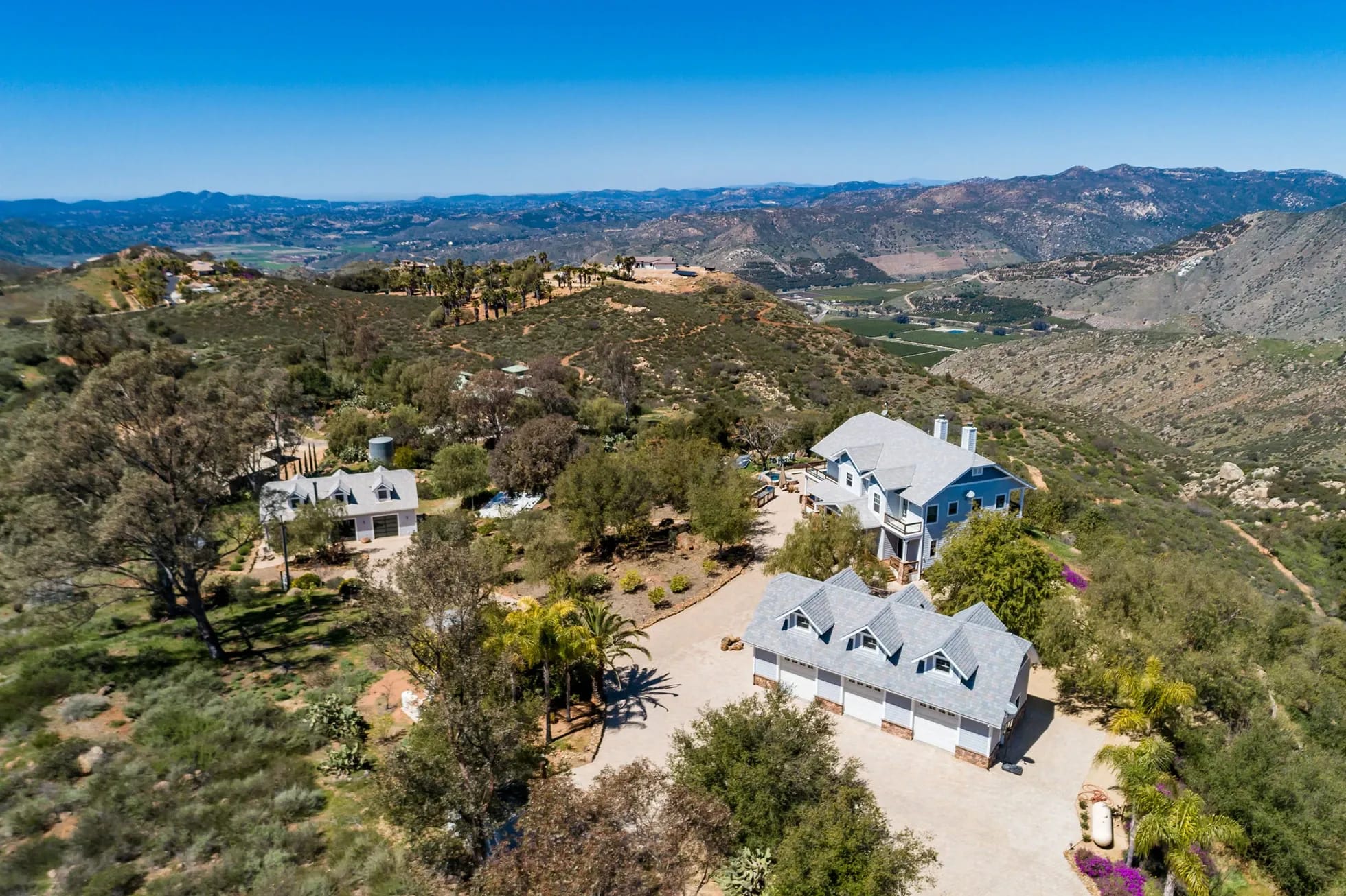Aerial view of a property featuring several houses nestled on a brush-covered hillside, with a valley and distant mountains under a clear blue sky.