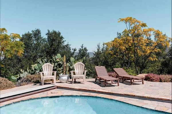 Outdoor swimming pool area with two Adirondack chairs and two lounge chairs on a stone patio, surrounded by trees and cacti under a clear blue sky.