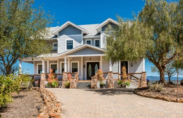 A light grey, two-story modern farmhouse-style house with a covered front porch, stone pillars, and a paved driveway. Large trees frame the house under a clear blue sky.