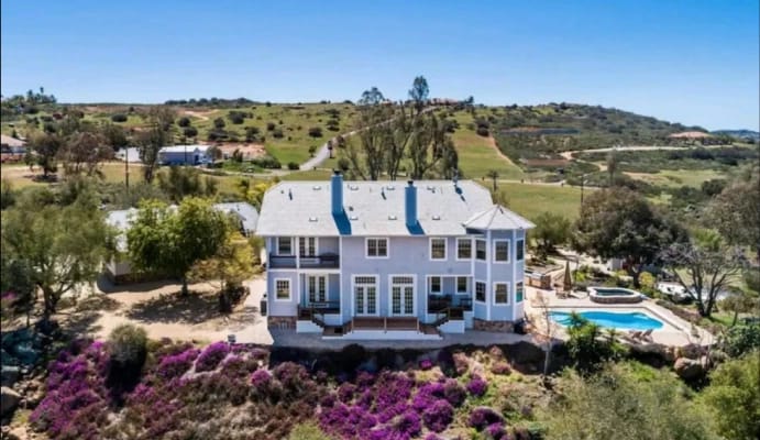 A large light blue house with a grey roof and multiple balconies, featuring a swimming pool and hot tub, set on a hill with purple flowers and rolling green hills in the background under a clear sky.