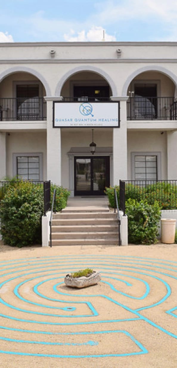 Exterior of Quasar Quantum Healing, a two-story white building with a sign above the entrance, and a blue labyrinth on the sandy ground in front.