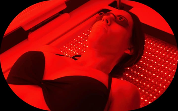 Woman lying in a red light therapy bed with protective eyewear, bathed in bright red light from LED panels.