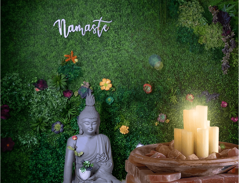 A peaceful setting with a Buddha statue in front of a green moss wall, accented by 'Namaste' and glowing candles on salt rocks.