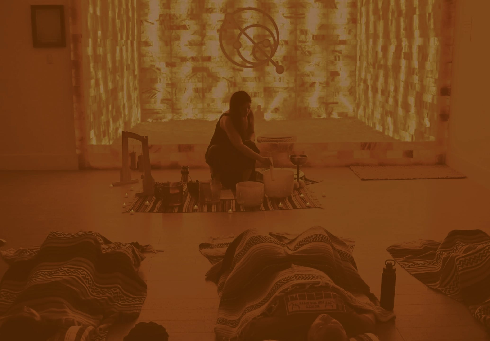 A person plays crystal bowls during a sound healing or meditation class, with participants lying under blankets in the foreground and an illuminated salt wall behind.