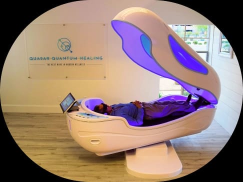 A person relaxes in a white, futuristic wellness pod with glowing purple interior lights, next to a 'Quasar Quantum Healing' sign.